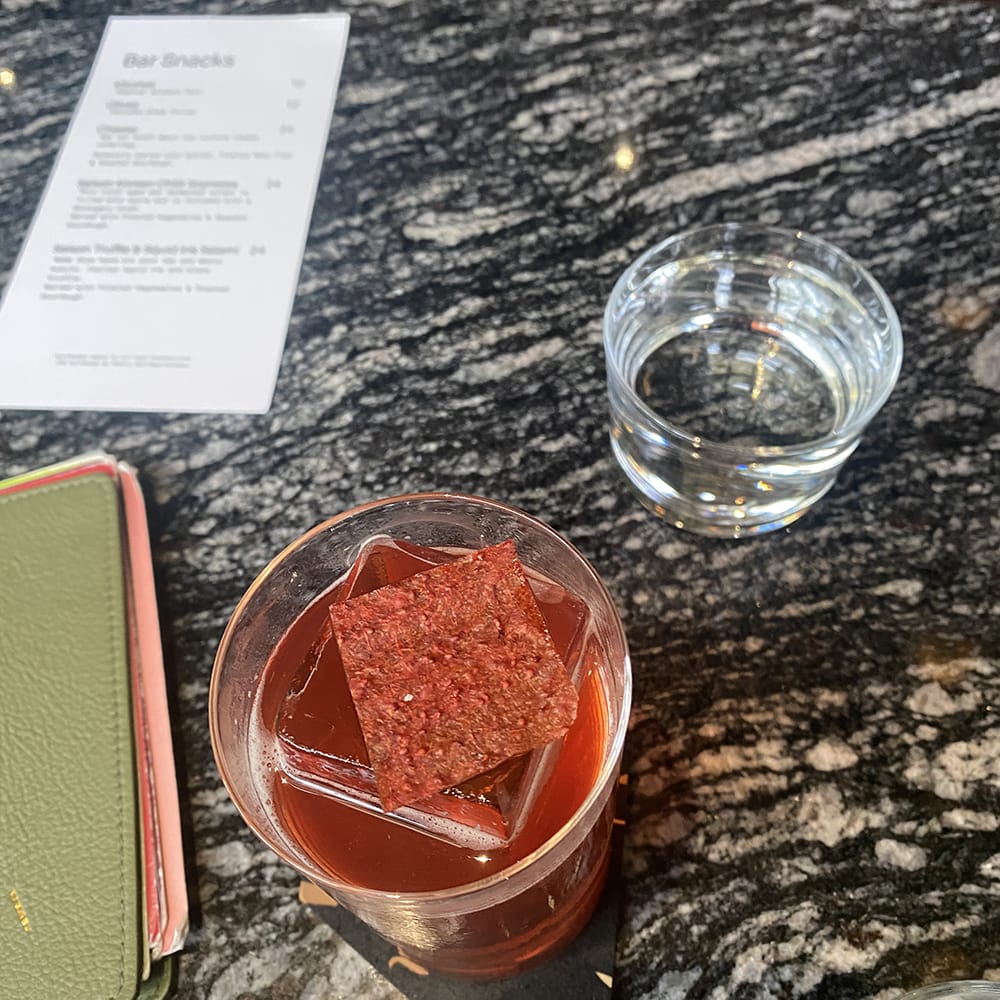 red cocktail with large block of ice and fruit leather on black marble table with green leather menu and glass of water